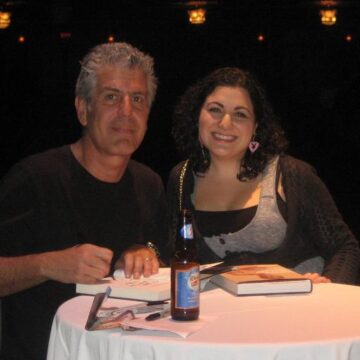 Anthony Bourdain posing with a female fan behind a table