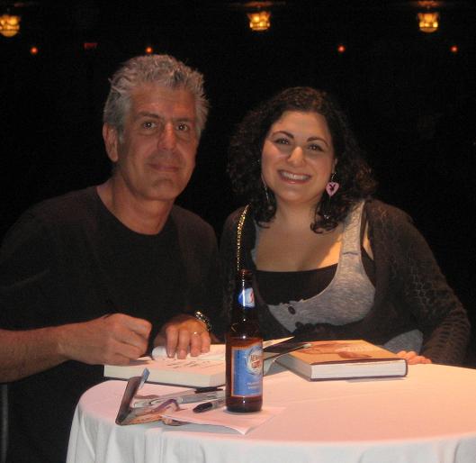 Anthony Bourdain posing with a female fan behind a table