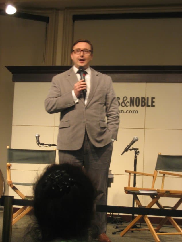 John Hodgman wearing a suit and tie standing in a room