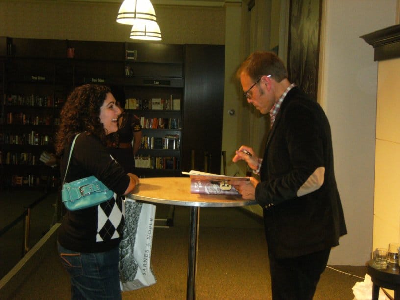 a woman standing at a table with Alton Brown who is signing a book