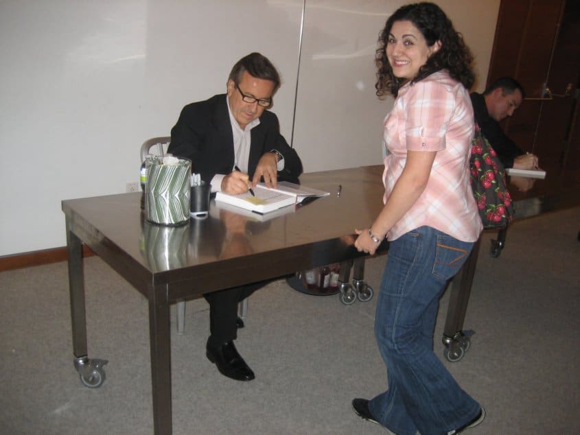 a woman posing by a table where Daniel Boulud is sitting and signing a book