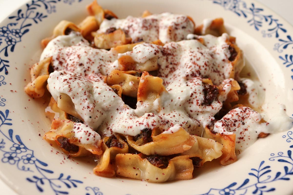 Armenian manti dumplings in a wide white bowl topped with yogurt sauce.