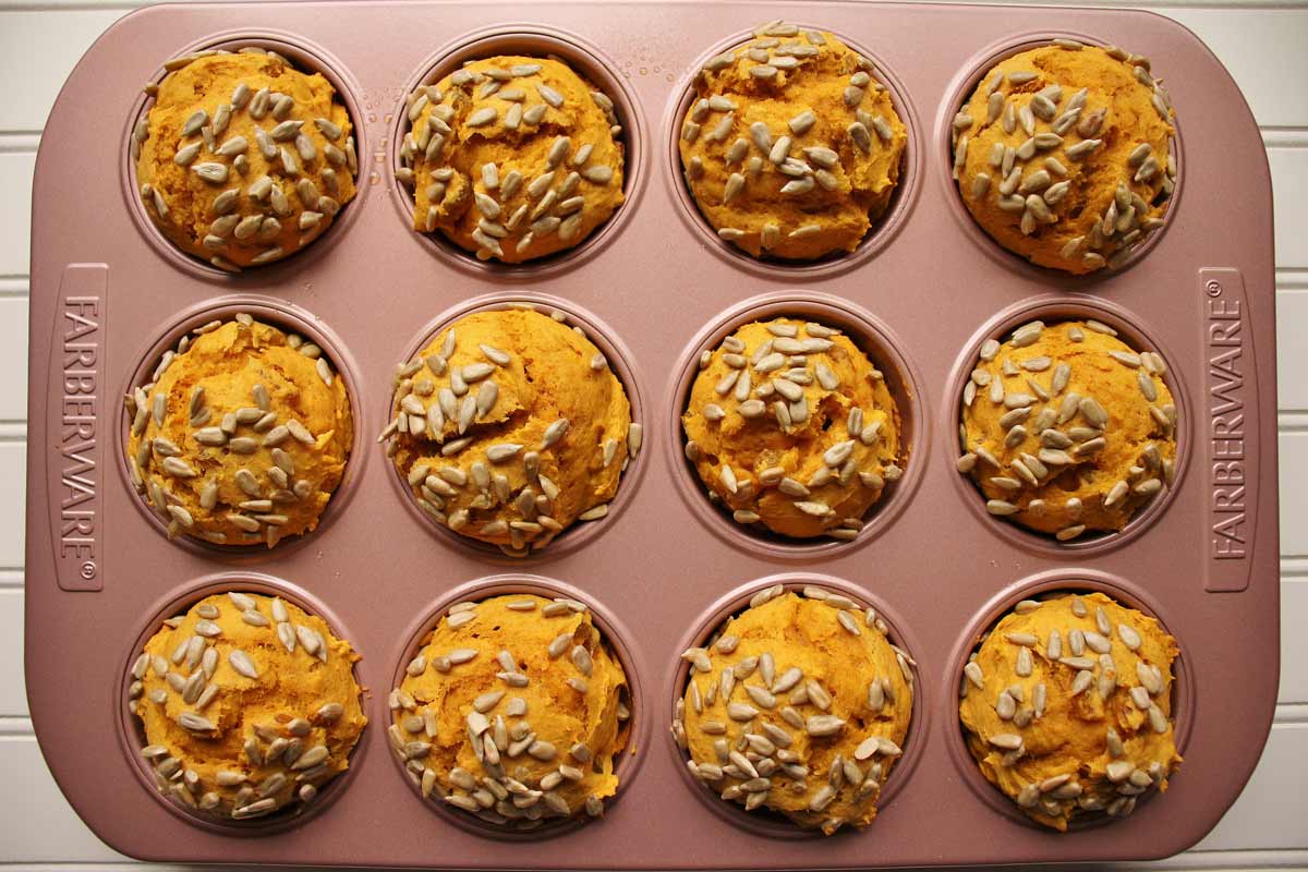 A pink muffin pan filled with bright orange muffins topped with sunflower seeds.