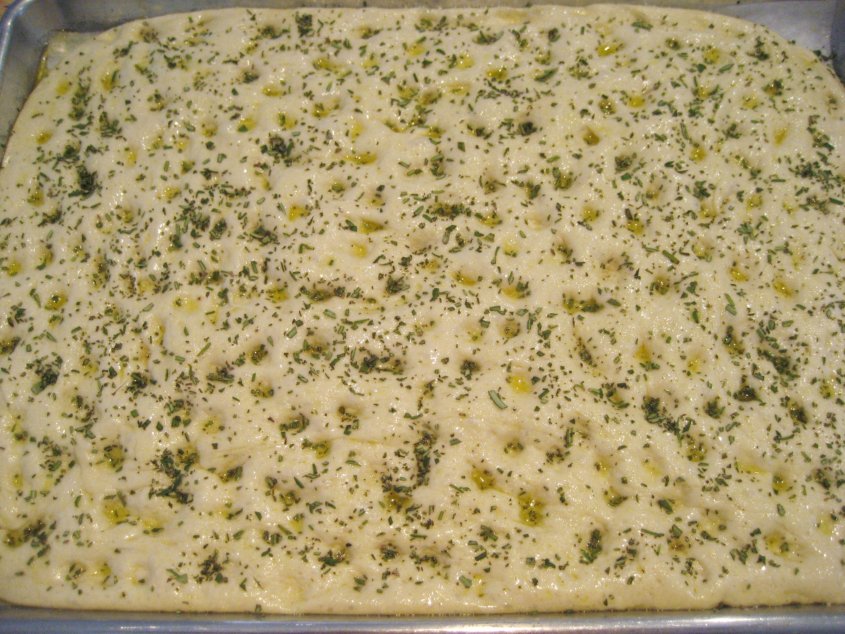 overhead view of focaccia dough spread out on a baking sheet topped with oil and herbs