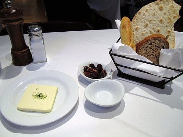 butter on a plate, a bowl of olives, and a bread basket on a white table