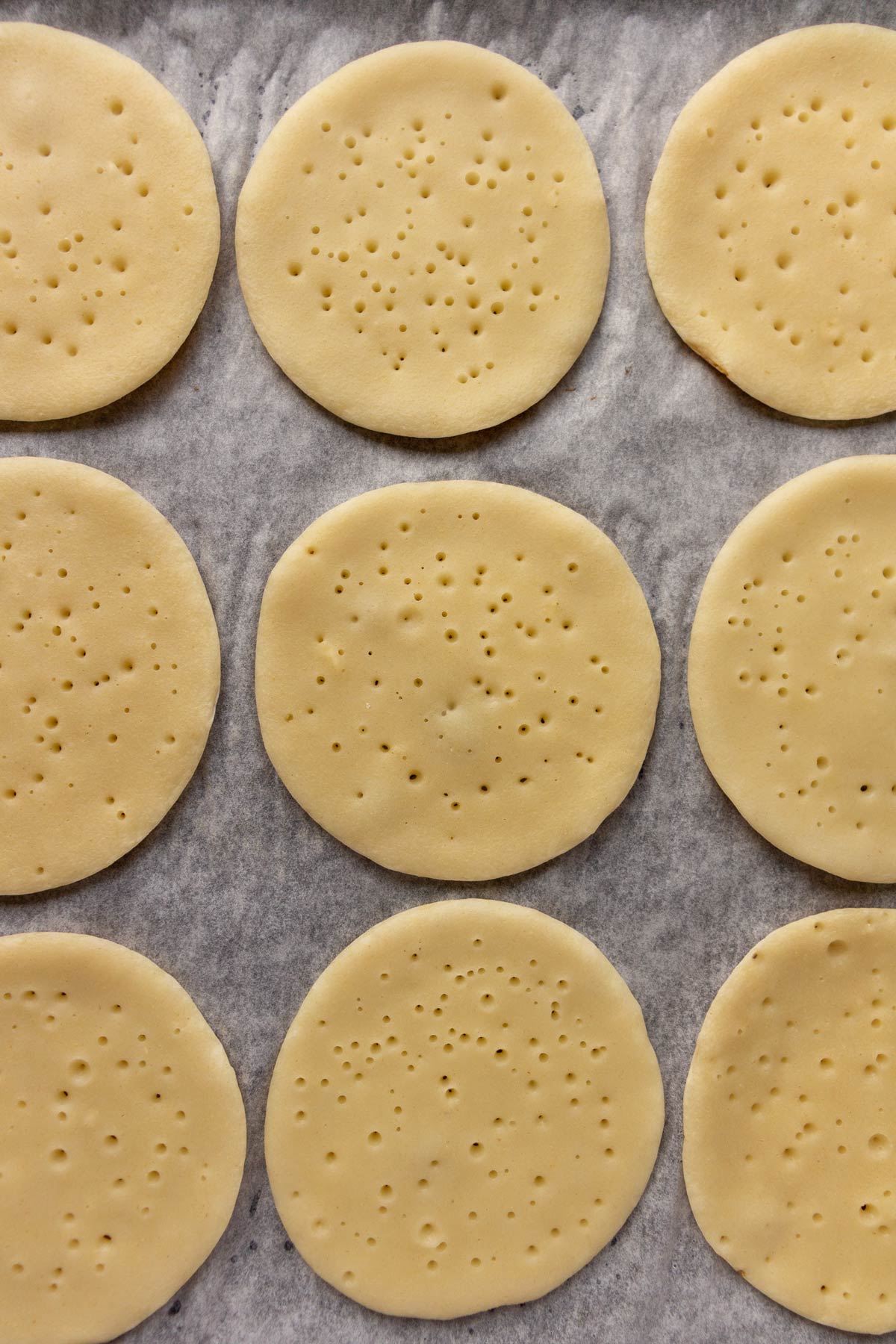 Rows of small pancakes with bubbles on the surface arranged in rows on parchment paper.