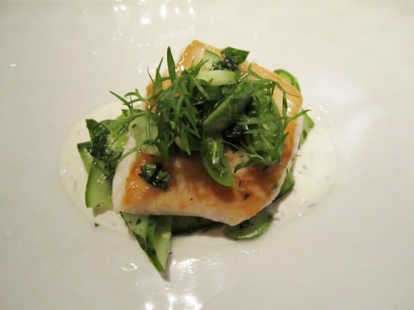 a slice of fish over cucumbers topped with green herbs on a white plate