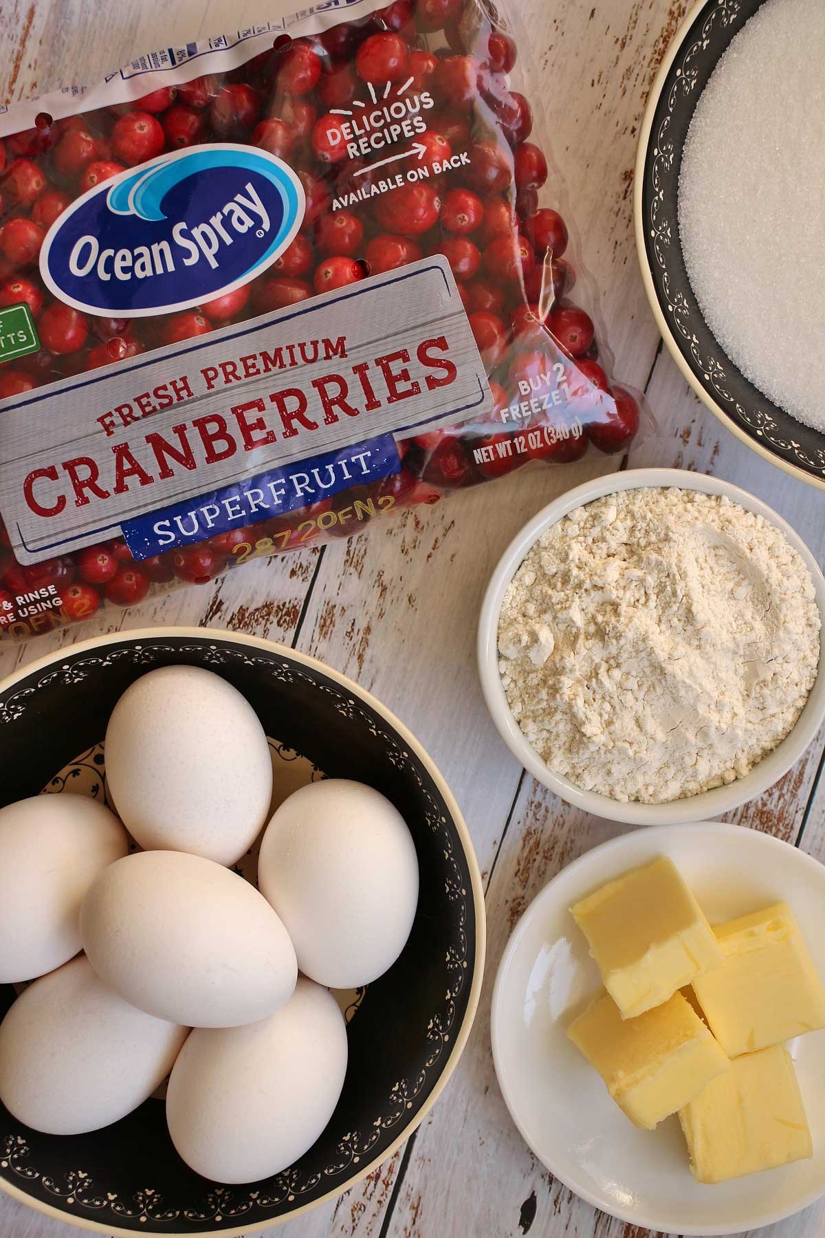 A bag of fresh cranberries, eggs, sugar, flour, and butter in dishes.
