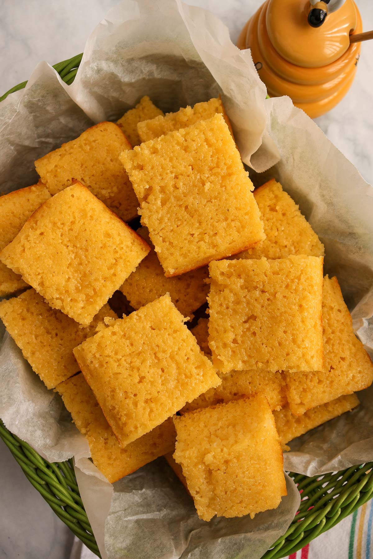 A green basket lined with parchment paper and filled with squares of cornbread piled high.