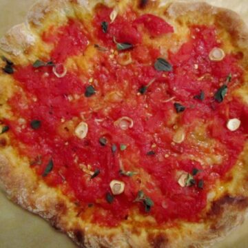 A closeup of a pizza with red tomato sauce, sliced garlic, and fresh herbs
