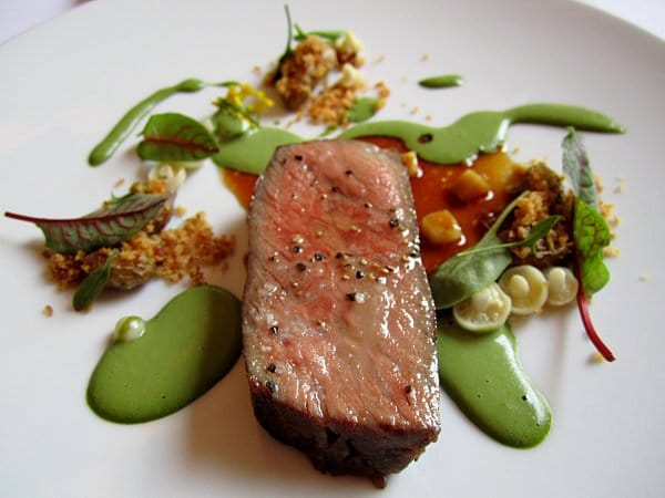 a slice of steak on a plate surrounded by drizzles of green sauce and tiny mushrooms