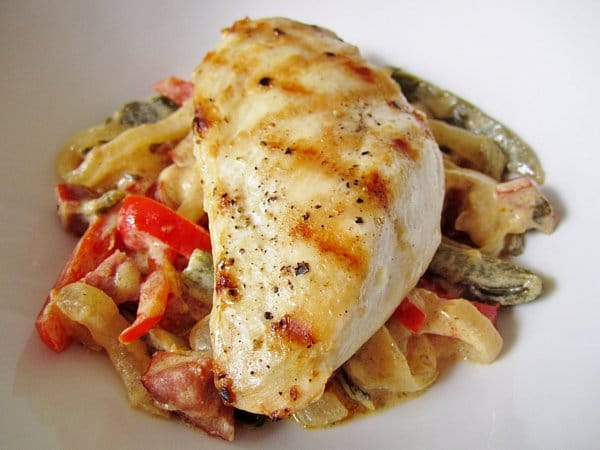 A closeup of a grilled chicken breast over cooked peppers and onions in a white dish