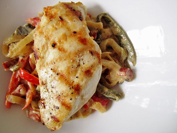 overhead closeup of a grilled chicken breast over cooked peppers and onions in a white dish