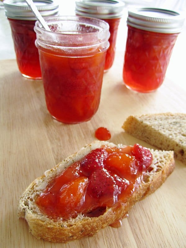 a slice of bread topped with strawberry peach preserves with jars of jam in the background