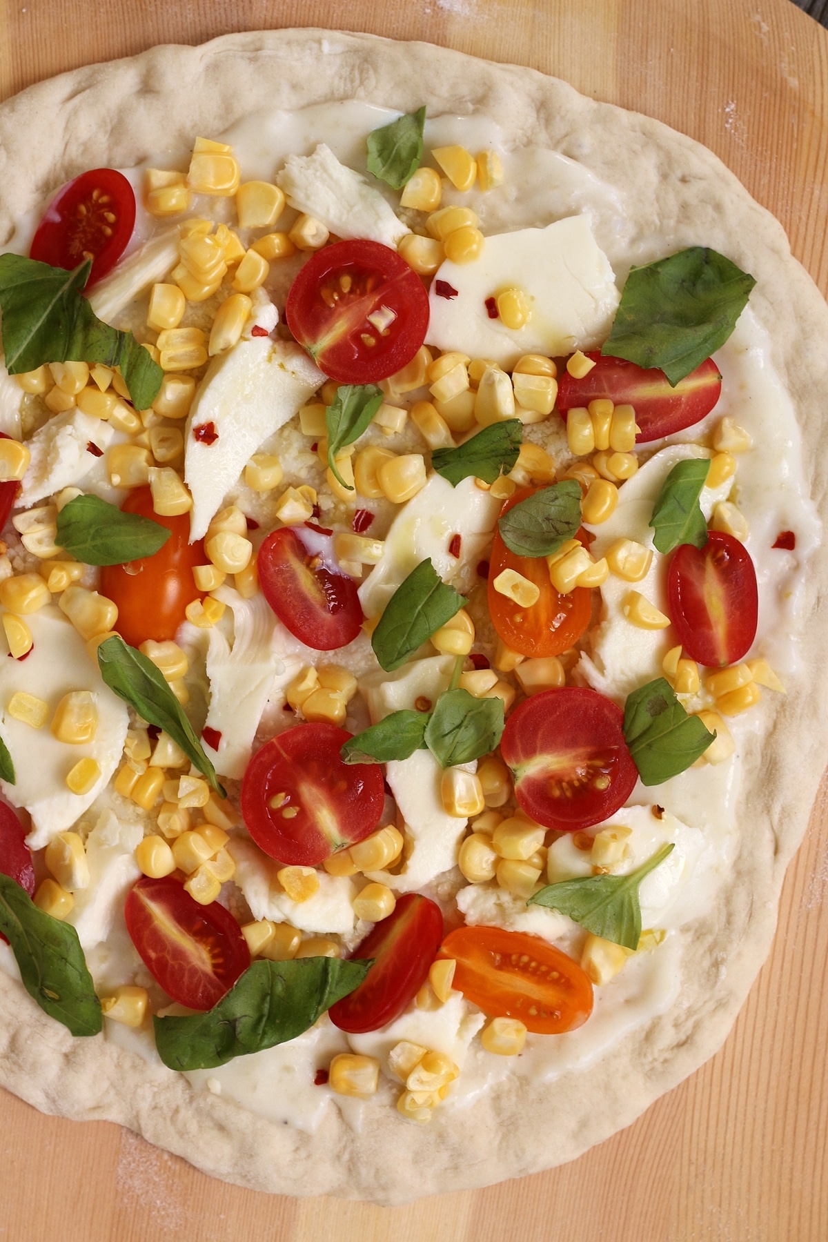 an unbaked pizza topped with corn, basil and cherry tomatoes on a wooden pizza peel