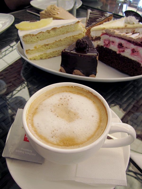 a cappuccino on a table with a platter of cake slices in the background