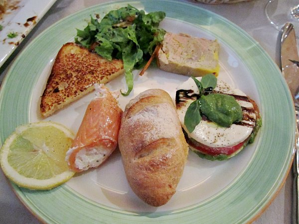 A plate of food on a table with bread, smoked salmon roll, pate, and mozzarella stack