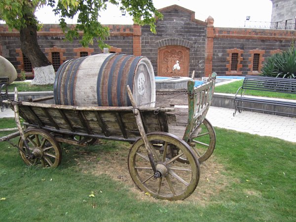 a wooden carriage topped with a large wooden barrel in front of a stone building