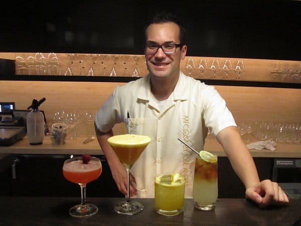 a man standing behind a bar lined with four different cocktails