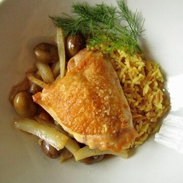 overhead view of a crispy chicken thigh with olives and rice in a white dish