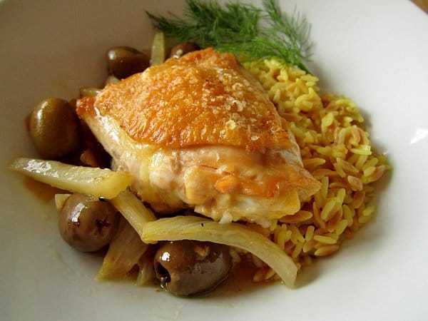 closeup of a crispy chicken thigh with olives, fennel, and rice pilaf in a white dish