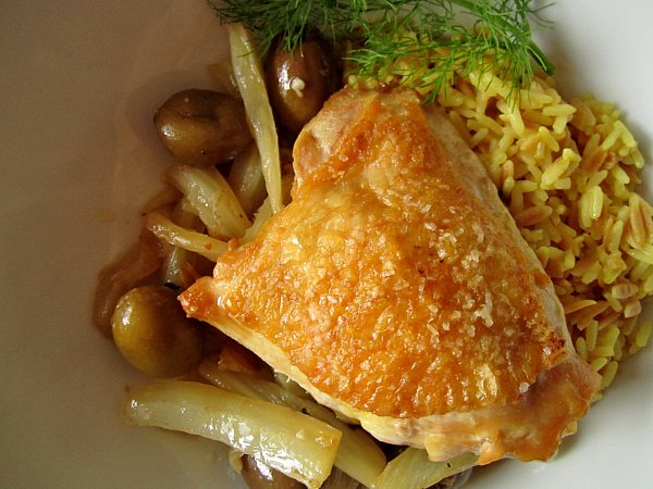 overhead closeup of a crispy chicken thigh with olives and rice in a white dish