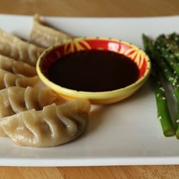 a bowl of sauce on a white plate with steamed dumplings and asparagus on either side