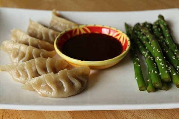 a bowl of sauce on a white plate with steamed dumplings and asparagus on either side