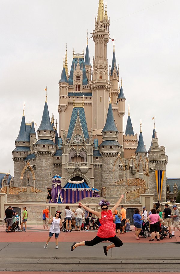 a woman in a red shirt jumping in front of Cinderella\'s Castle
