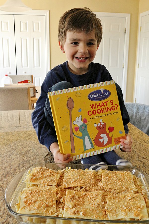 a boy holding a cookbook while sitting on the counter behind a casserole dish