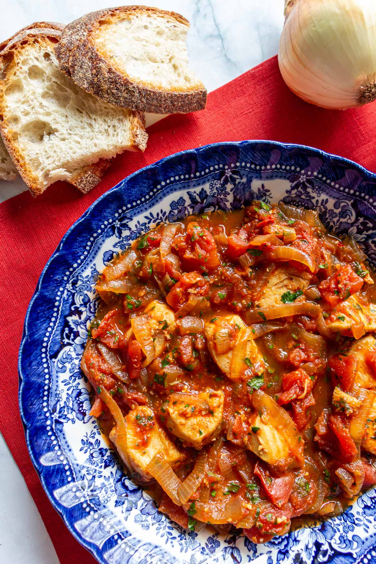 Closeup of a bowl of chicken stew with tomatoes and onions with sliced bread next to it.