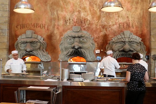 people preparing food in front of three large pizza ovens with faces