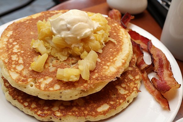 a stack of pancakes topped with pineapple pieces and butter with bacon in the side