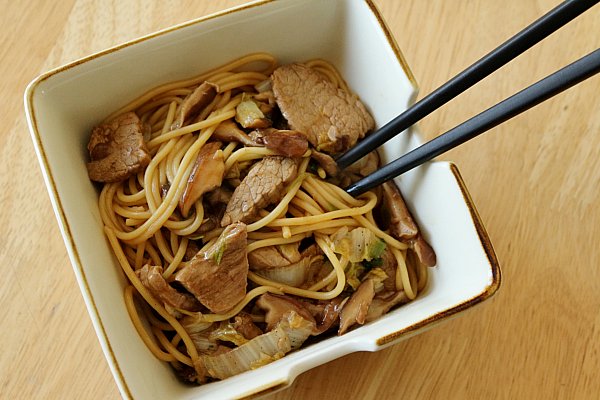 overhead view of a square bowl filled with Asian noodles with sliced pork, mushrooms, and chopsticks