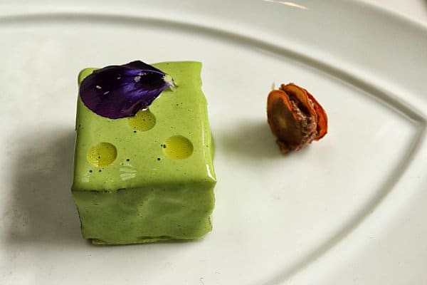 a small square bite of food on a plate with a purple flower petal on top