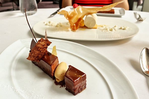 a chocolate dessert on a white plate with another dessert in the background