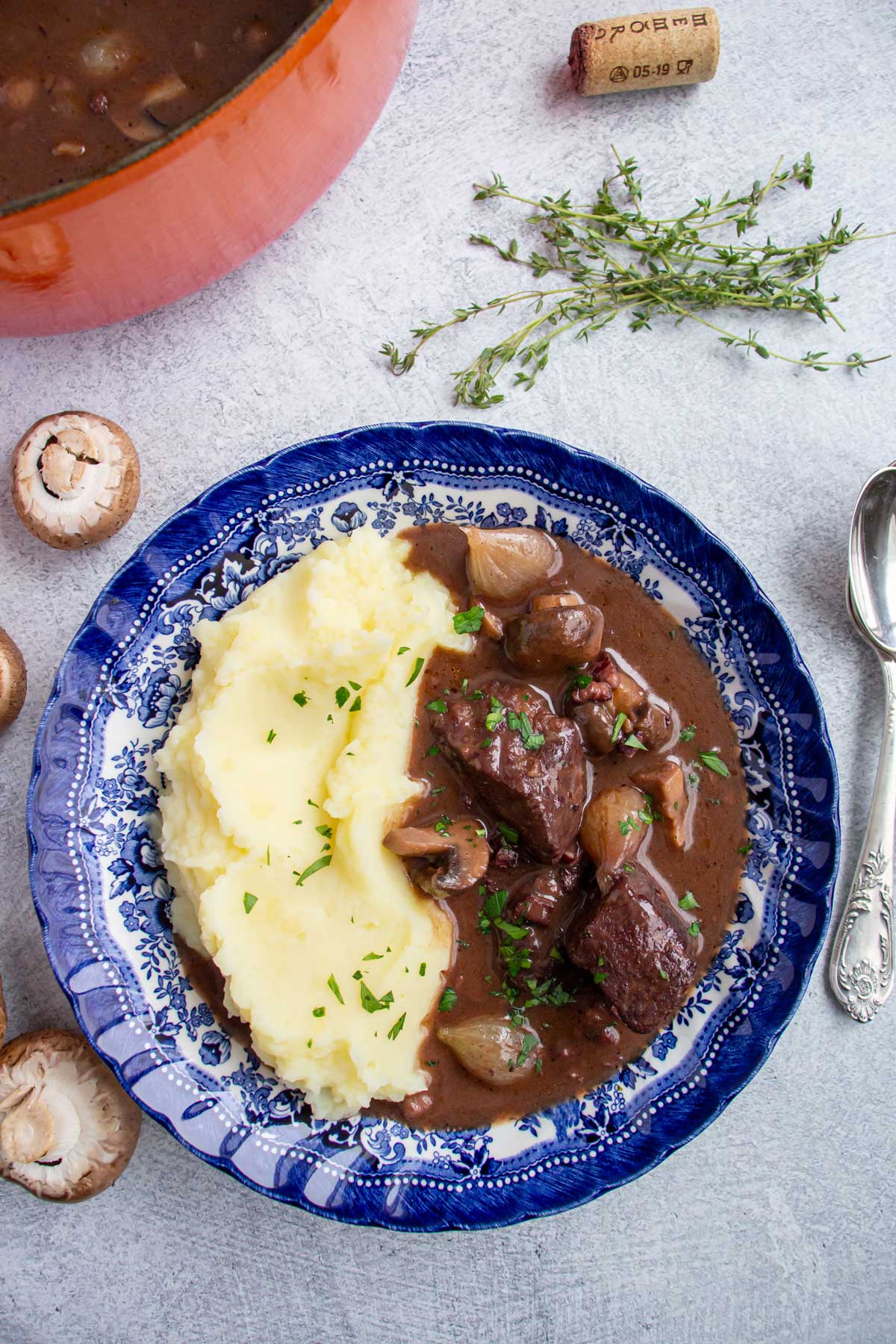 A bowl of beef burgundy stew with mashed potatoes, next to mushrooms and springs of thyme.