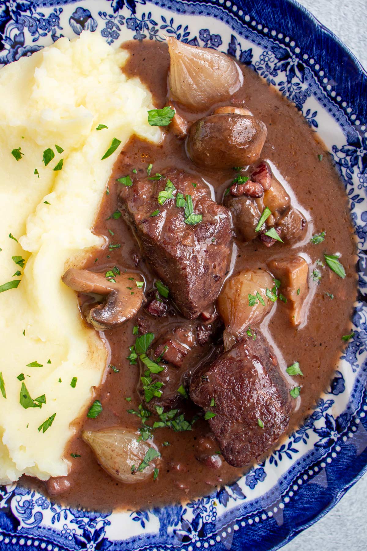 Burgundy beef stew with mushrooms and pearl onions served with mashed potatoes in a blue bowl.