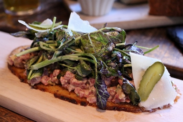 a thin bread toast topped with chopped meat and greens on a wooden board