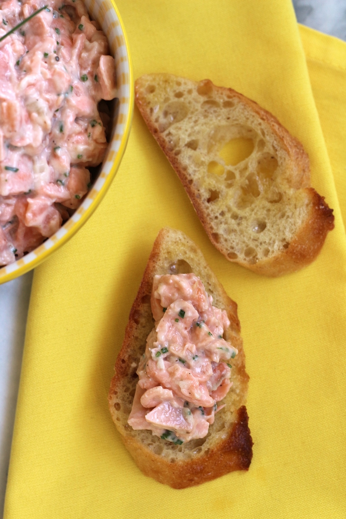 2 toasted baguette slices on a yellow napkin, 1 topped with salmon rillettes spread