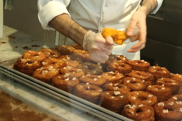 a closeup of a man\'s hands piping filling into fried doughnuts on a tray