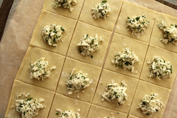 Assembling cheese boreks: puff pastry cut into squares, topped with grated feta cheese and parsley