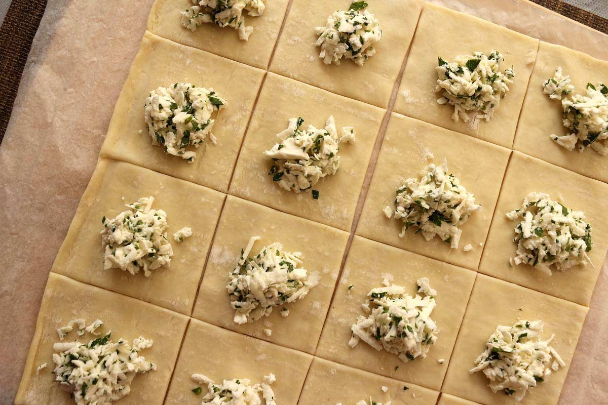 Assembling cheese boreks: puff pastry cut into squares, topped with grated feta cheese and parsley.