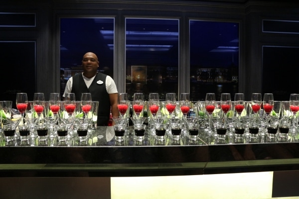 rows of glasses of drinks lined up on a bar with a man standing behind it