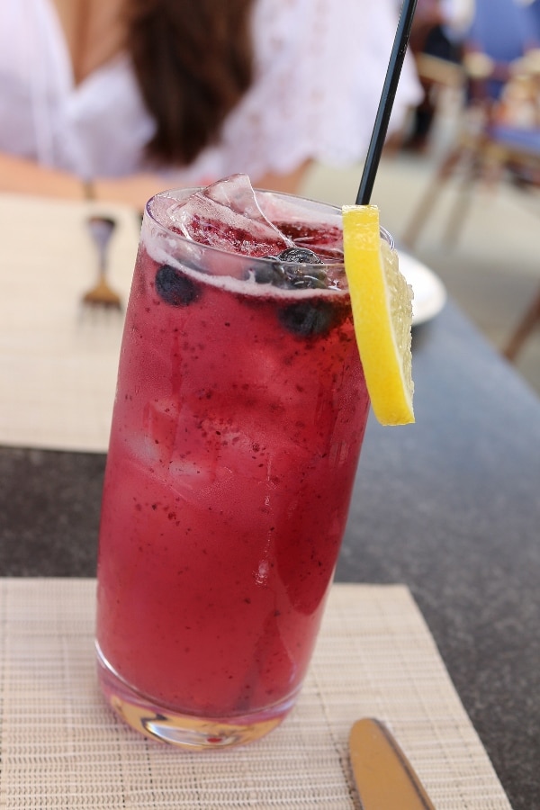 a tall glass filled with a bluish drink topped with floating blueberries and a lemon slice