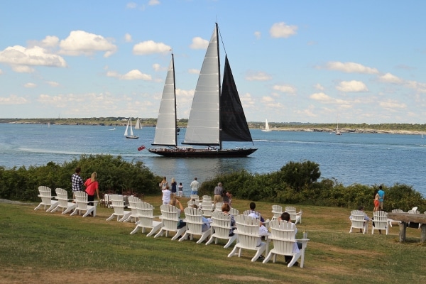 a boat with large black and white sails in the water by people in Adirondack chairs