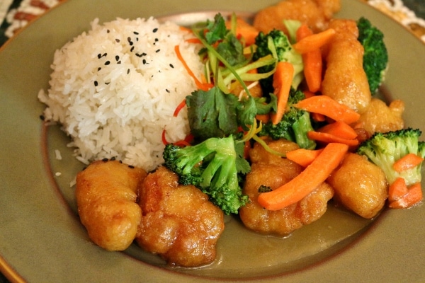 A plate of crispy honey chicken with broccoli and carrots, and a scoop of white rice