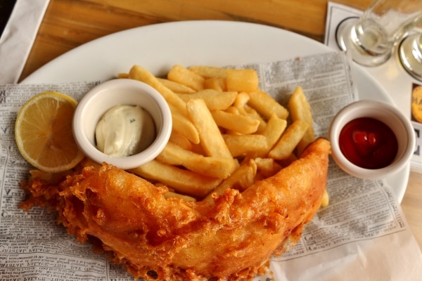 A plate of fish and chips on fake newspaper with cups of tartar sauce and ketchup