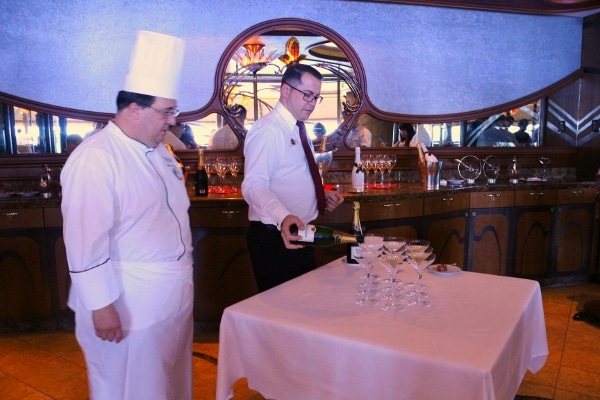 a man standing at a table next to a chef pouring glasses of Champagne