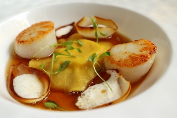 side view of a big ravioli in broth served with seared scallops and charred onions